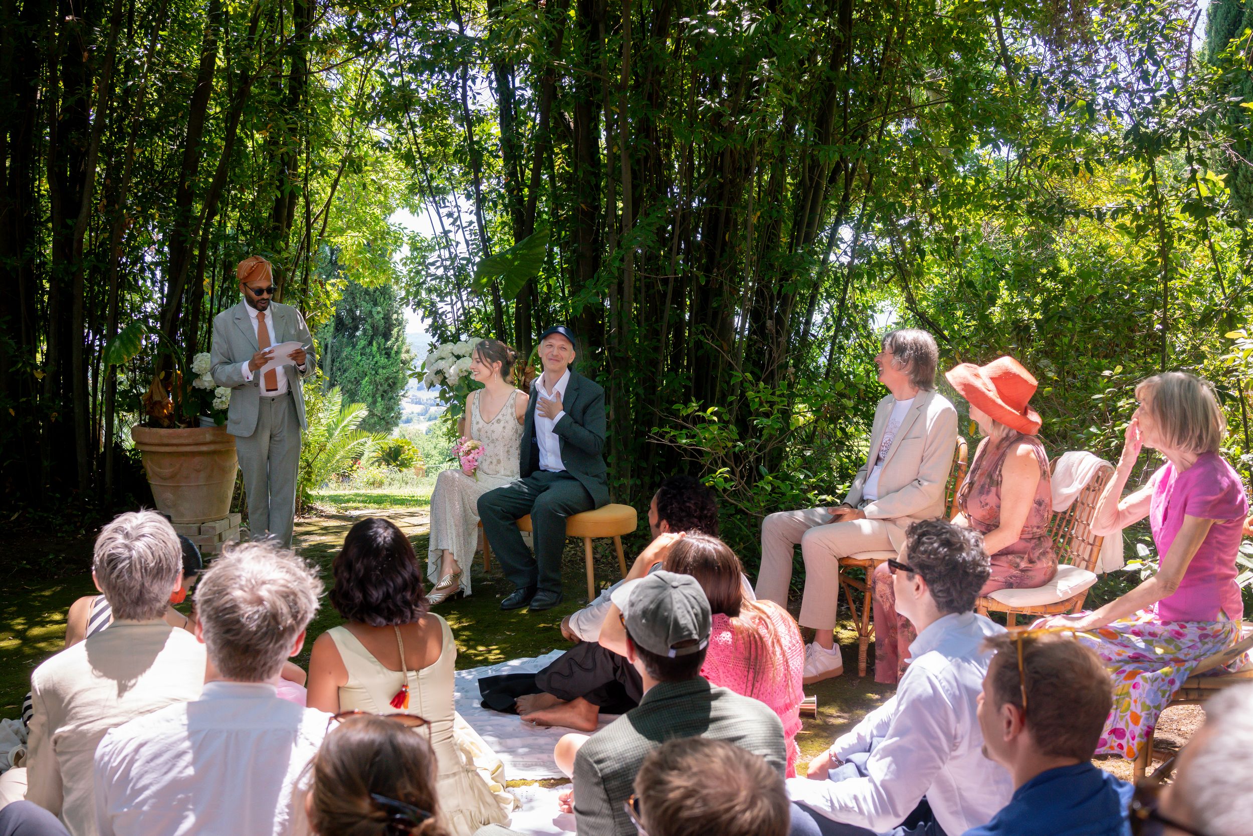 Outdoor gathering with people seated watching a small ceremony in a lush bamboo garden on a sunny day.