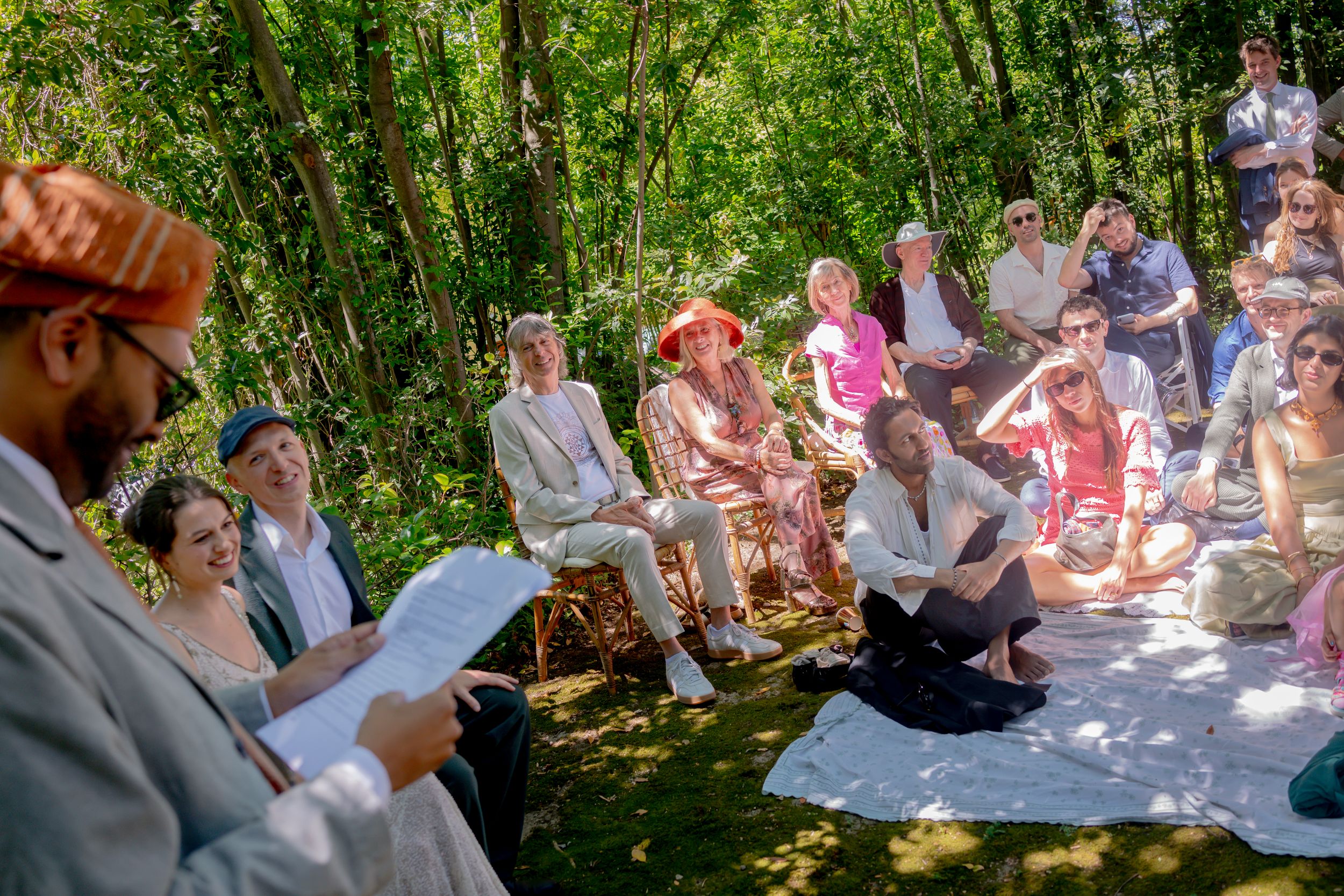 Outdoor wedding ceremony in a woodland setting with guests seated on chairs and blankets in bright summer sunshine.