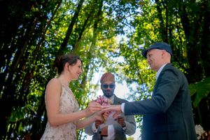 Couple exchanging rings during an outdoor wedding ceremony surrounded by lush green trees with officiant present.