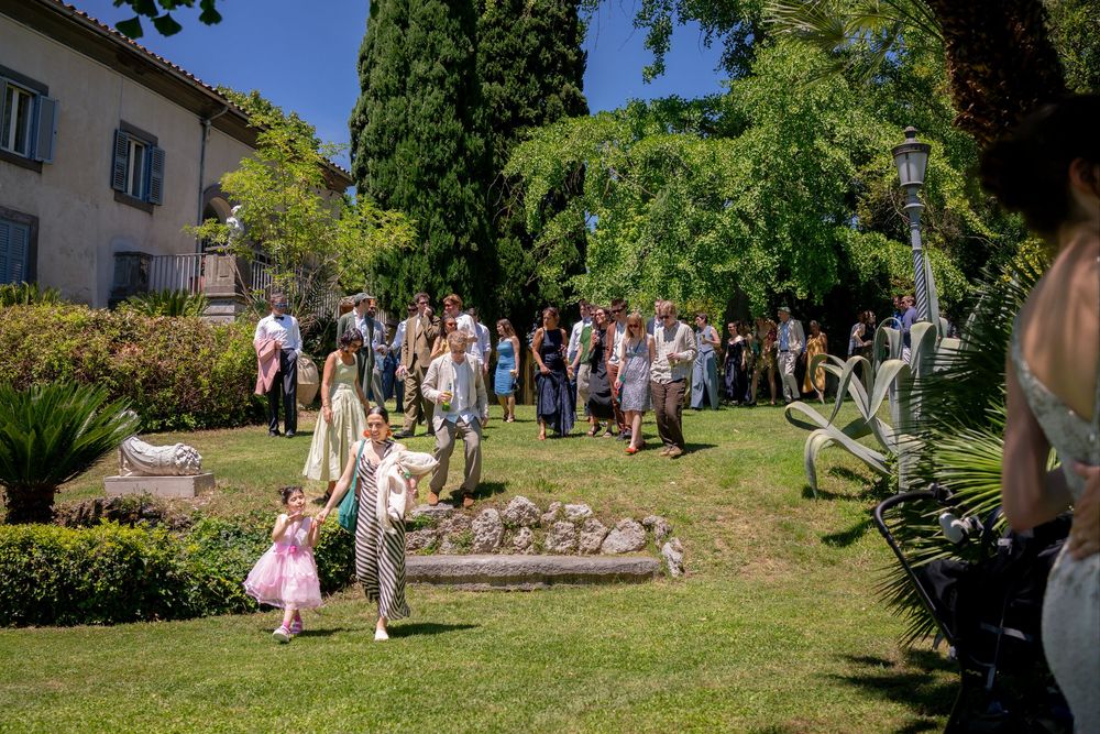 Guests mingle in a sunny garden at an outdoor wedding reception, with a historic villa and lush trees in the background.