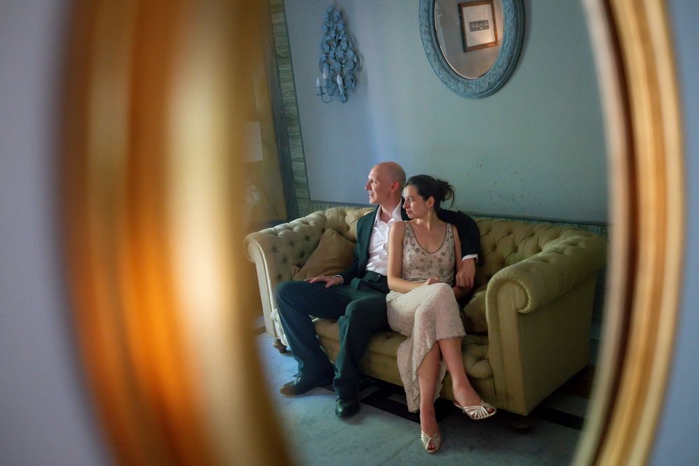 A couple sits intimately on a velvet sofa, reflected in a vintage oval mirror in an elegant blue room.