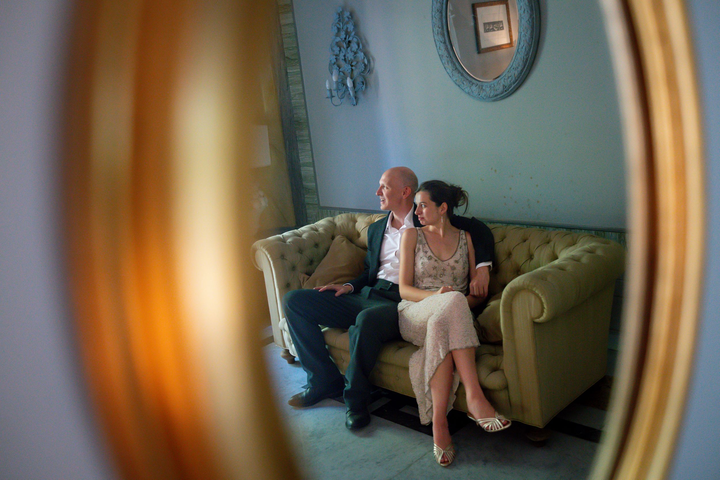 A couple sits intimately on a velvet sofa, reflected in a vintage oval mirror in an elegant blue room.