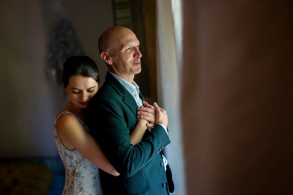 A bride embraces a bald groom from behind near a window, both holding hands in a tender wedding moment.
