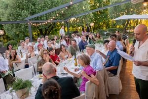 Wedding reception dinner outdoors with guests seated at decorated tables while a man gives a toast.