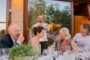 Guests laughing and enjoying speeches at an outdoor wedding reception dinner with string lights.