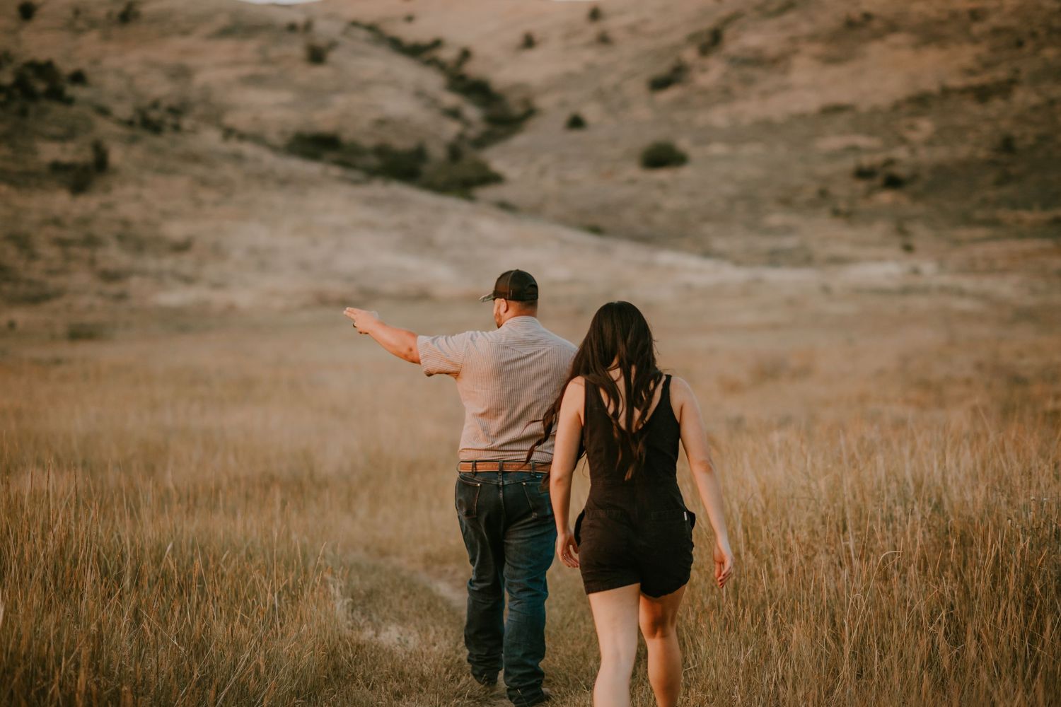 Couple walking through golden dry grass fields with rolling hills, man pointing into distance, woman in black dress.
