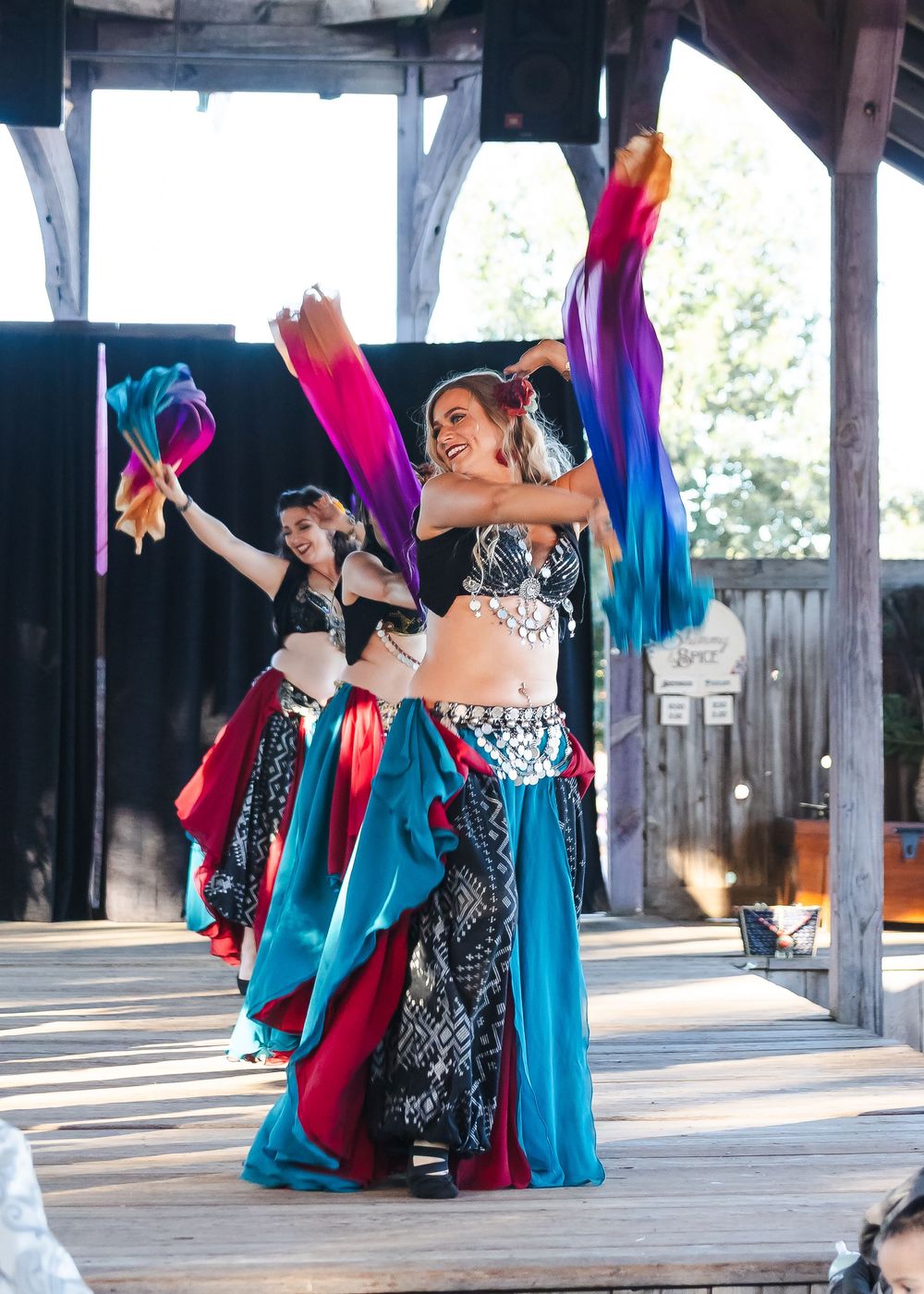 Two belly dancers in colorful teal and black costumes wave rainbow silk fans on an outdoor stage.