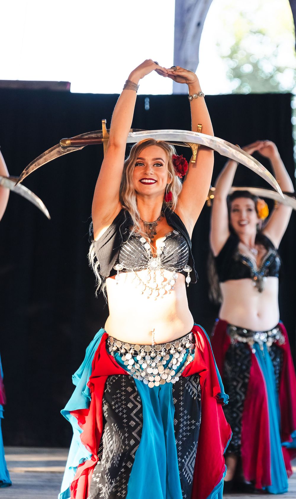 Belly dancer in colorful costume balances swords overhead while performing on stage with other dancers behind her.