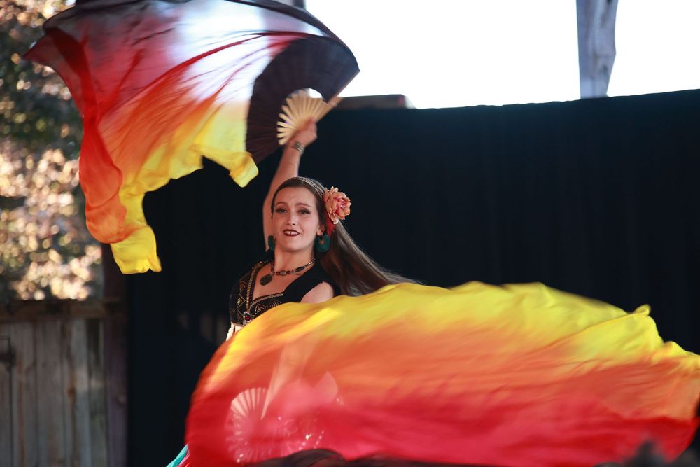 Woman performing belly dance with vibrant red, orange, and yellow silk fan veils outdoors in a black costume.