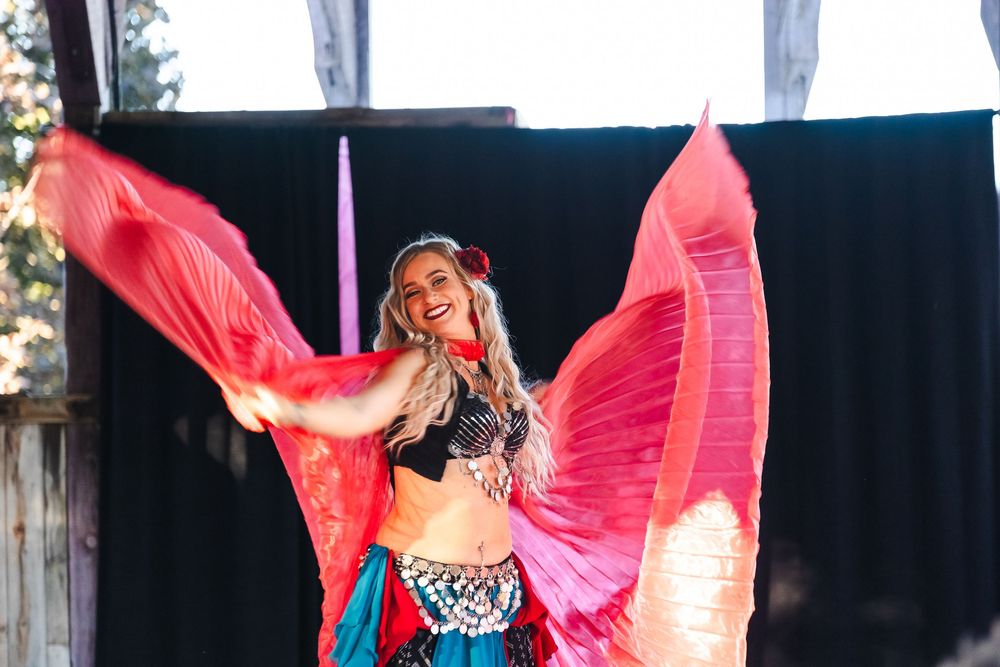 Belly dancer in colorful costume with red isis wings smiling outdoors at festival performance.