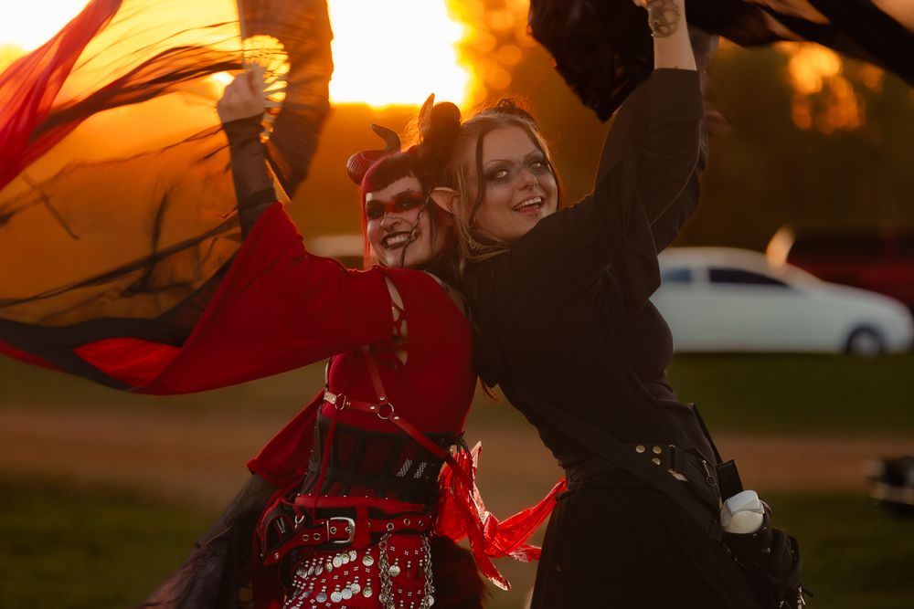 Two women in red and black costumes dance outdoors at sunset, waving colorful fabric in a festive performance.