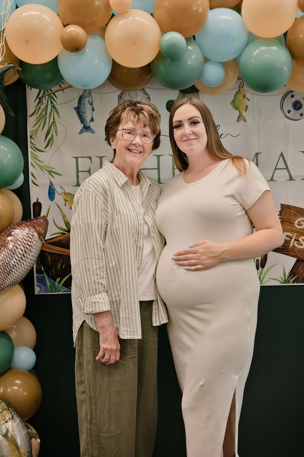 Two women pose at a fishing-themed baby shower with teal and tan balloon arch backdrop.