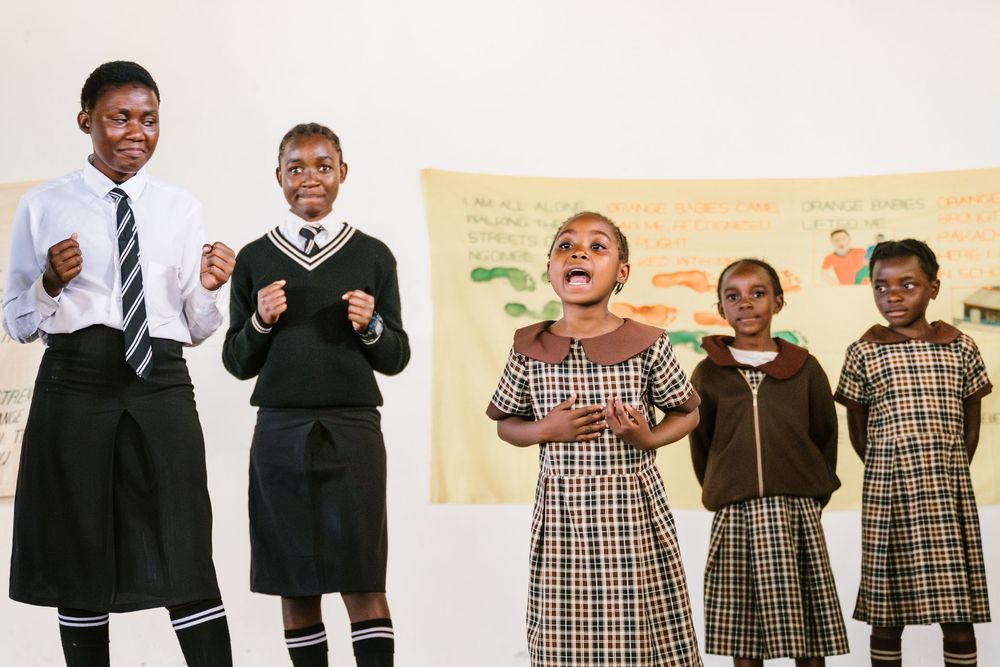 Five African schoolgirls in uniforms perform a lively skit in front of a colorful poster in a bright classroom.