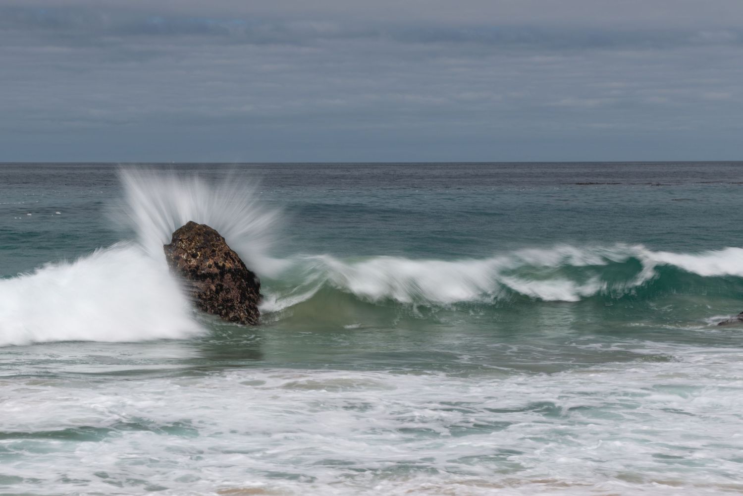 Ocean waves crash dramatically against a large rock on a sandy beach, creating a spectacular white water splash.