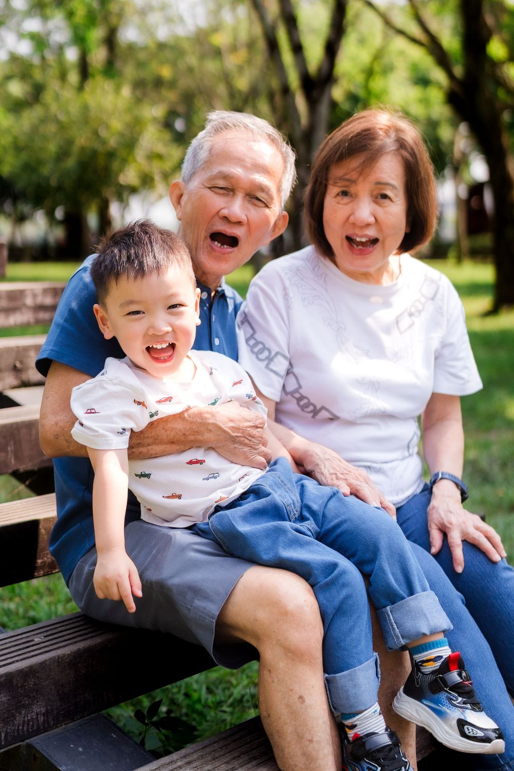 Grandparents laughing with toddler grandson on park bench outdoors.