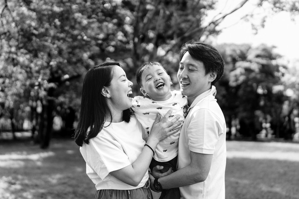 Black and white photo of happy Asian family with parents holding laughing toddler in a sunny park.