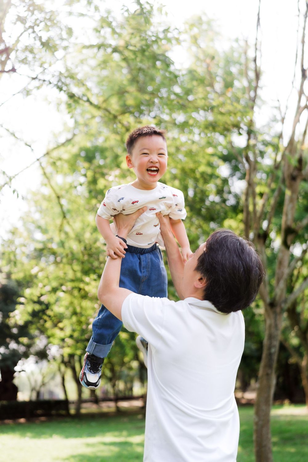 A father joyfully lifts his laughing young son high in the air outdoors in a green park.