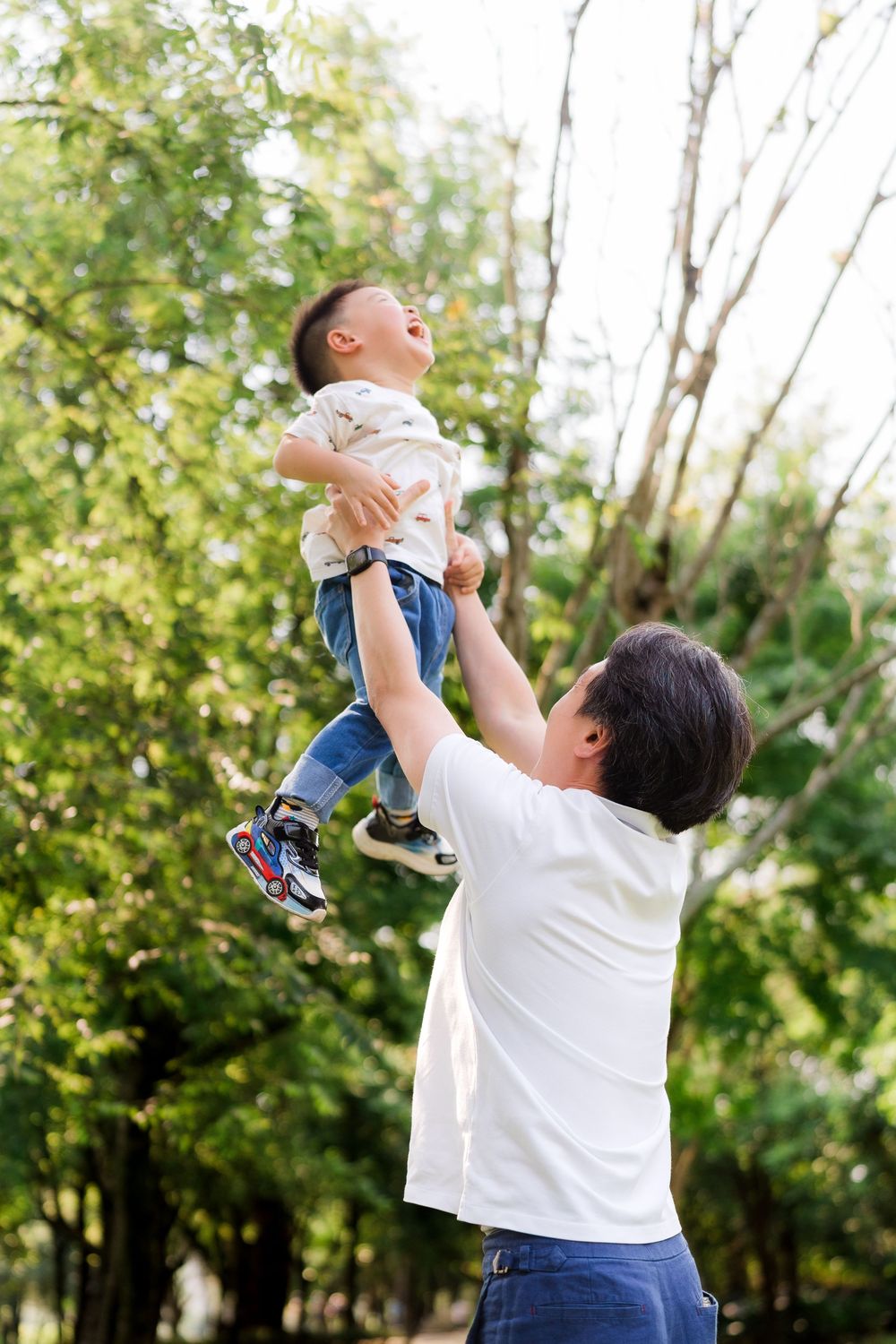 Father joyfully lifts laughing toddler son high in the air outdoors in a lush green park.