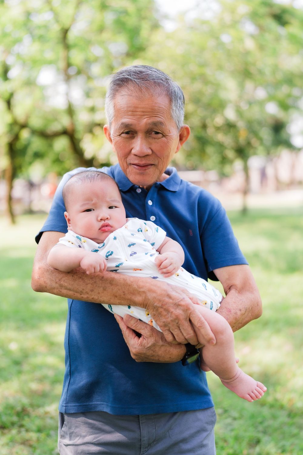 Elderly Asian grandfather tenderly holding newborn baby outdoors in green park setting.