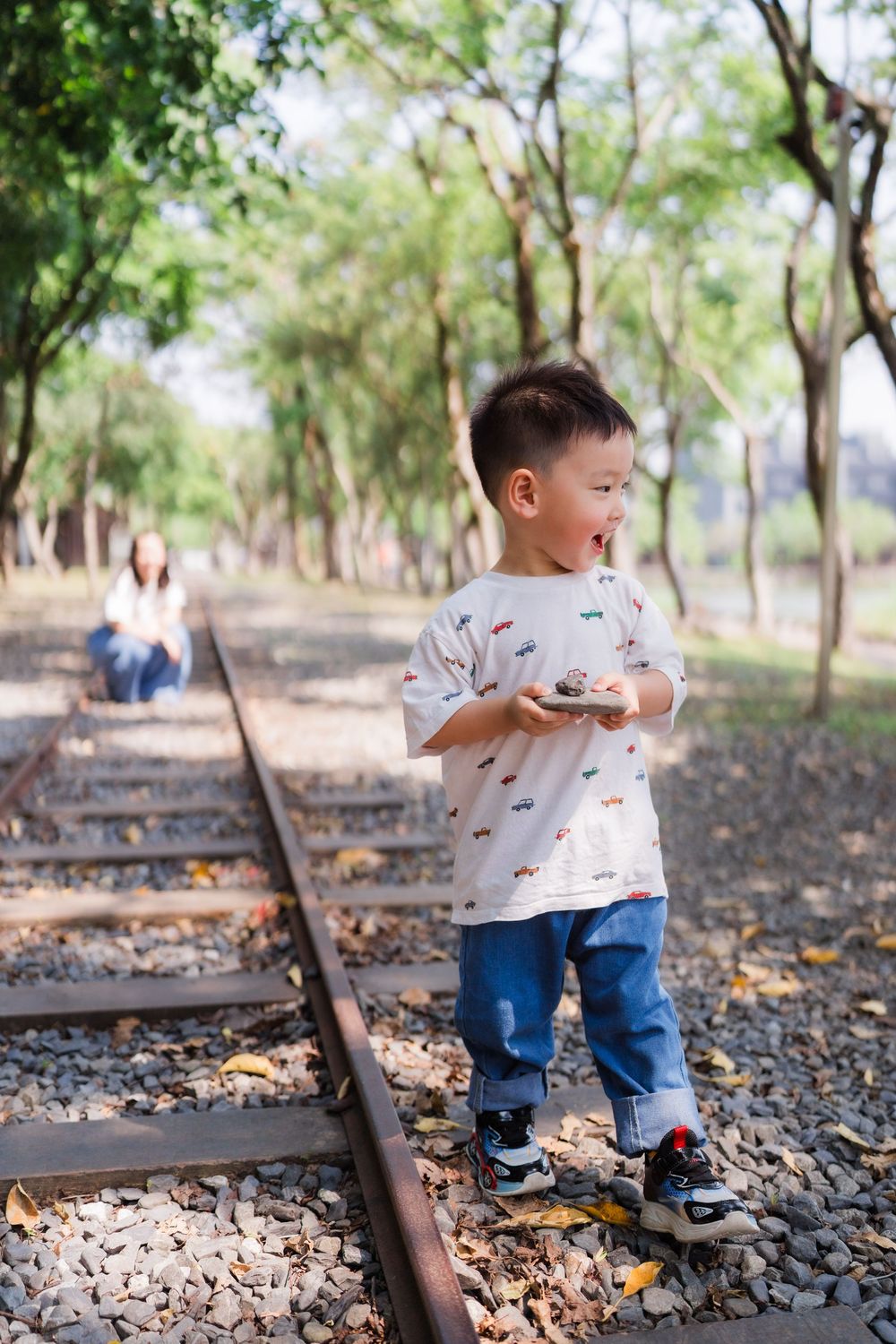 A young Asian boy stands on railway tracks holding a small rock while his mother sits behind him.