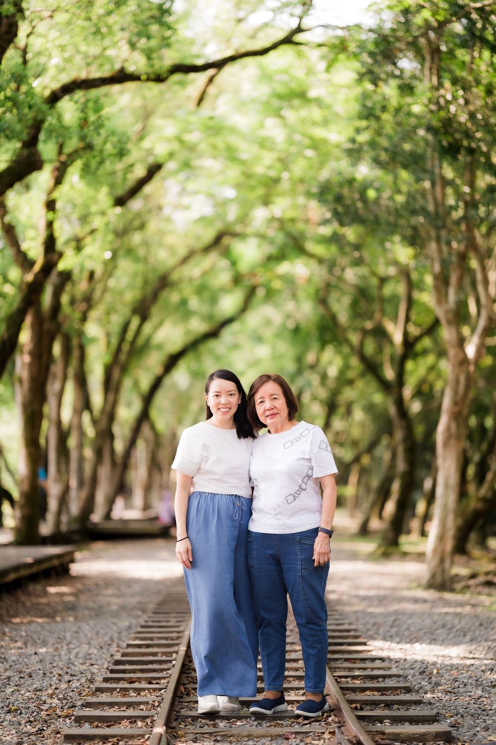 Two Asian women in white tops and blue pants pose together on railway tracks lined with green trees.