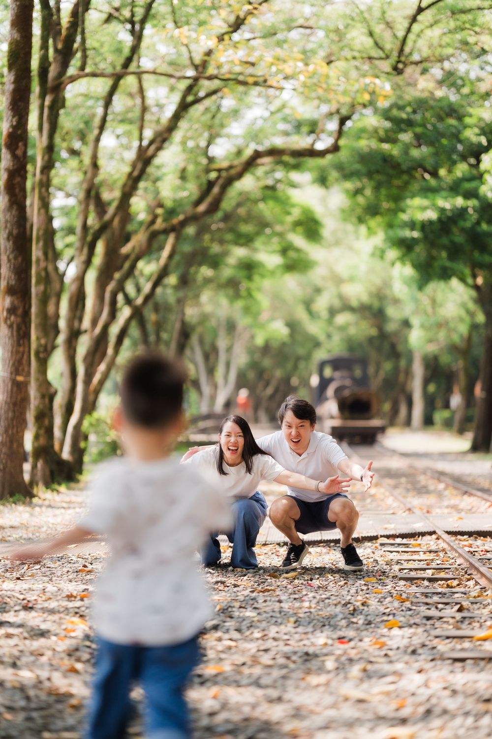 Excited Asian parents crouching with open arms as child runs toward them on railway.