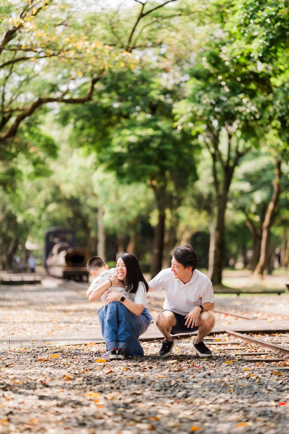 Asian couple crouches on old railway tracks in a lush green park, sharing a warm, candid moment.