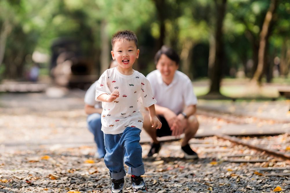 A joyful young Asian boy runs toward the camera on a leaf-covered path as his father watches.