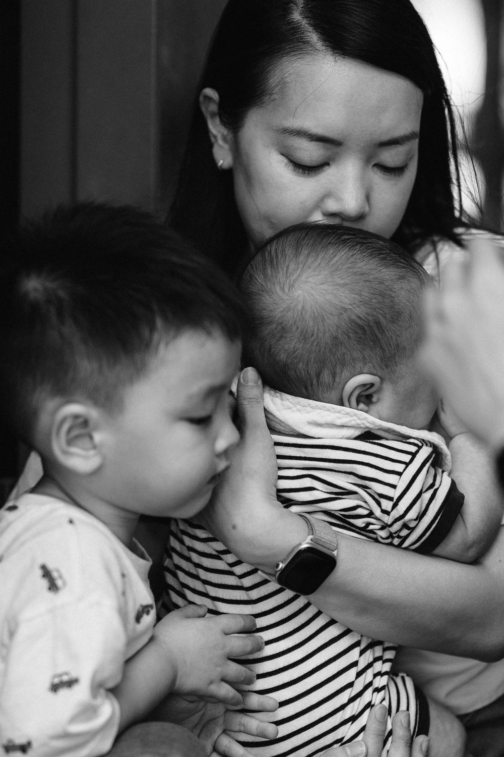 A black and white photo of an Asian mother kissing her baby while her toddler son hugs the infant.