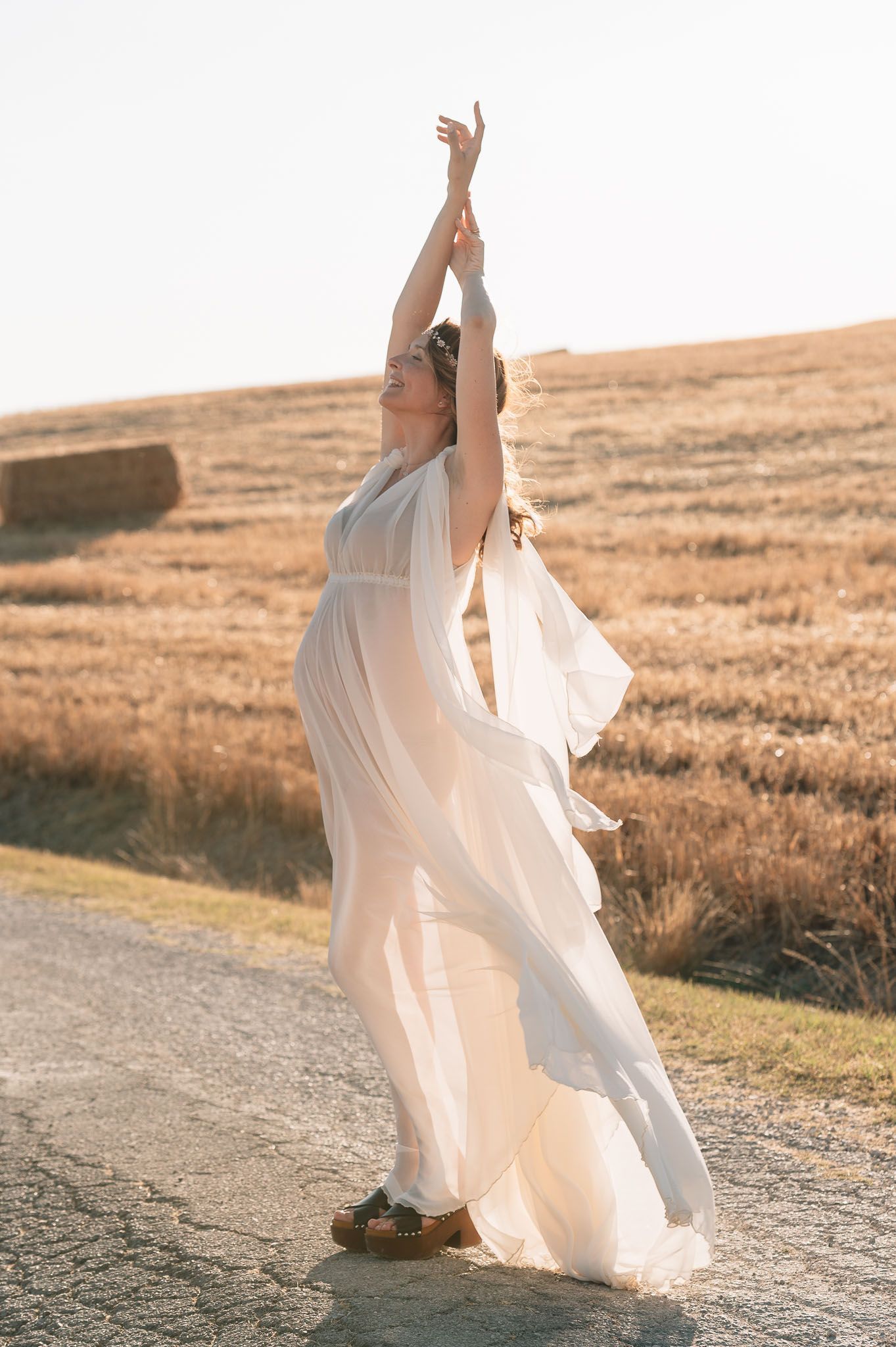 Woman in flowing white dress dancing gracefully on a rural road beside golden wheat fields under bright sunlight.