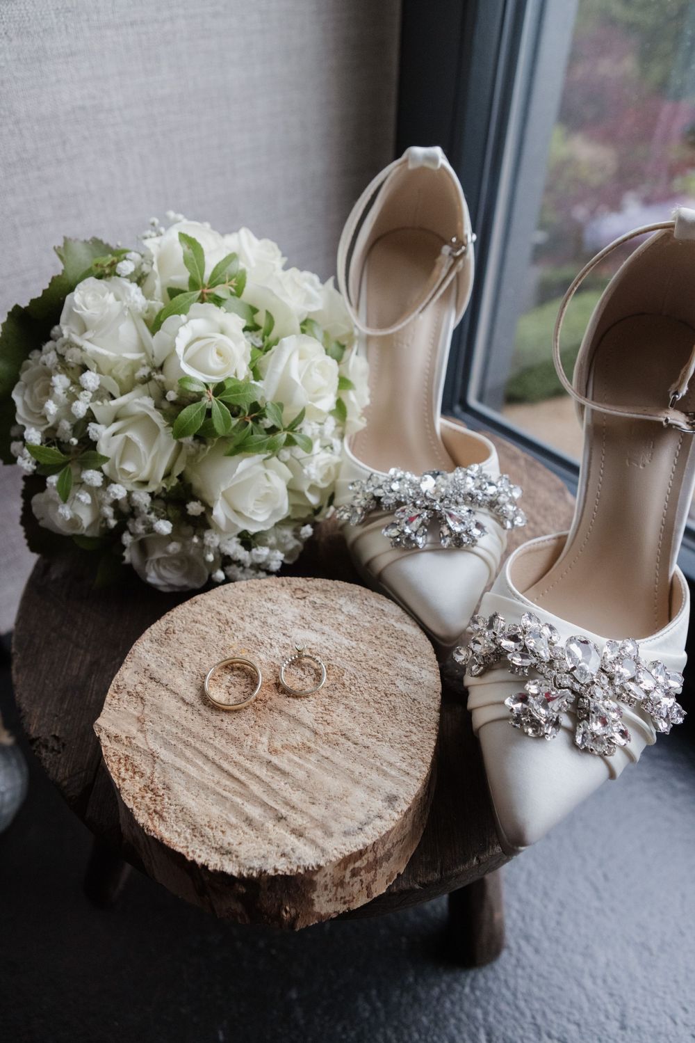 Eheringe und funkelnde Brautschuhe mit weißem Rosenstrauß auf Holzscheibe – Hochzeitsdetails in Sonsbeck am Niederrhein.