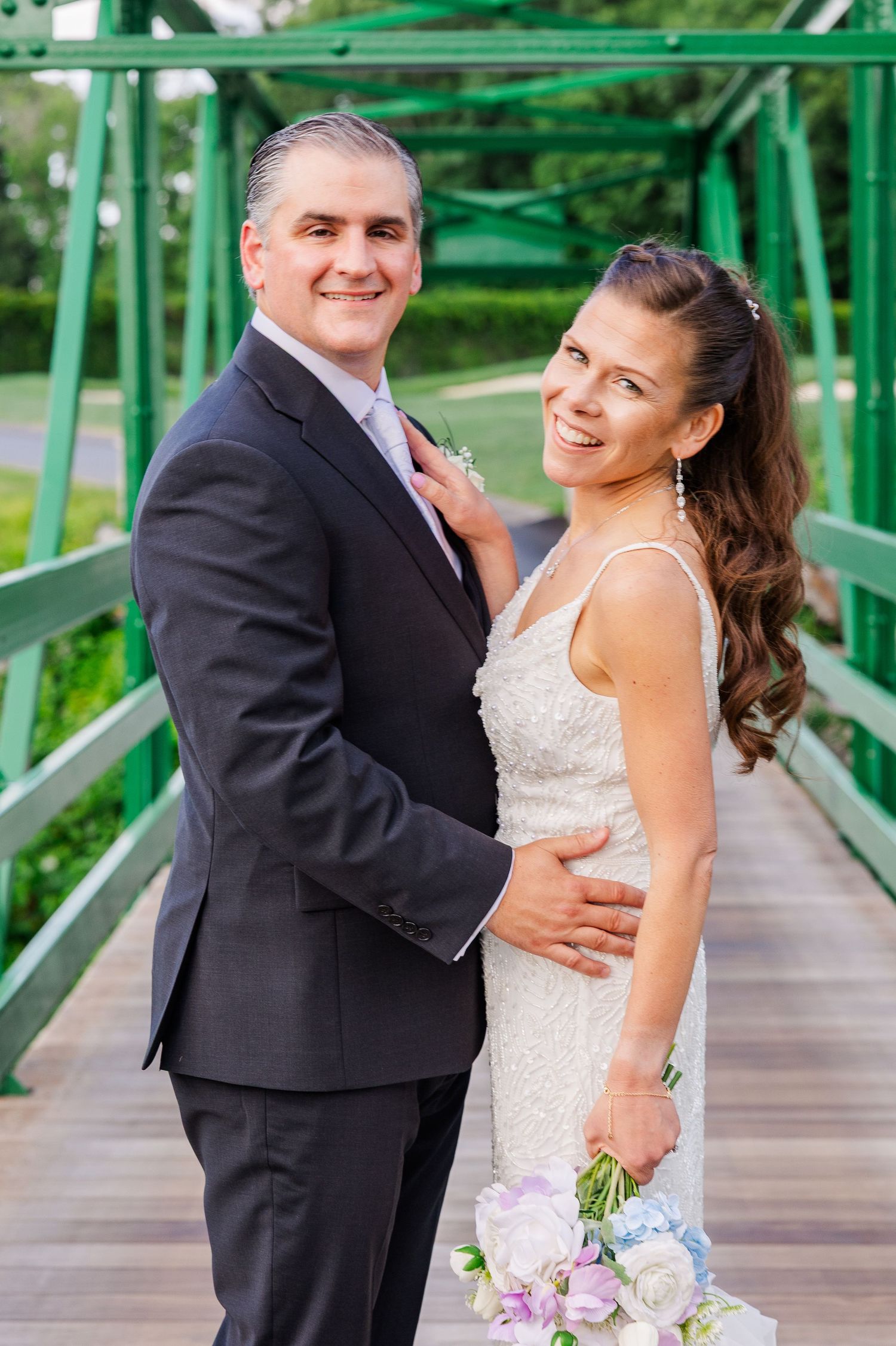 A smiling bride and groom pose on a green bridge, she holds a pastel bouquet in a white gown, he wears a dark suit.