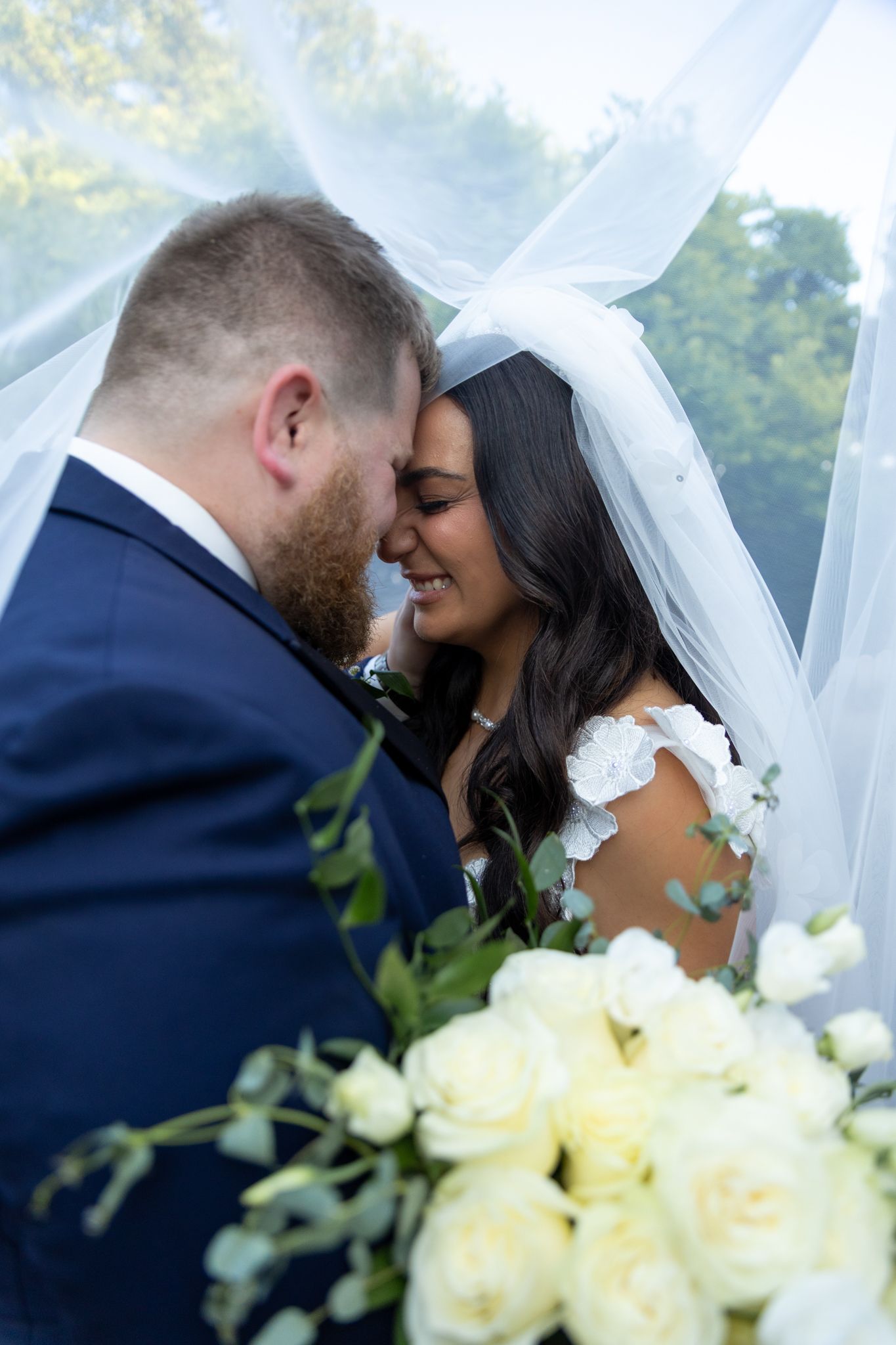 Bride and groom share a romantic moment under a flowing veil, holding white roses bouquet on their wedding day.