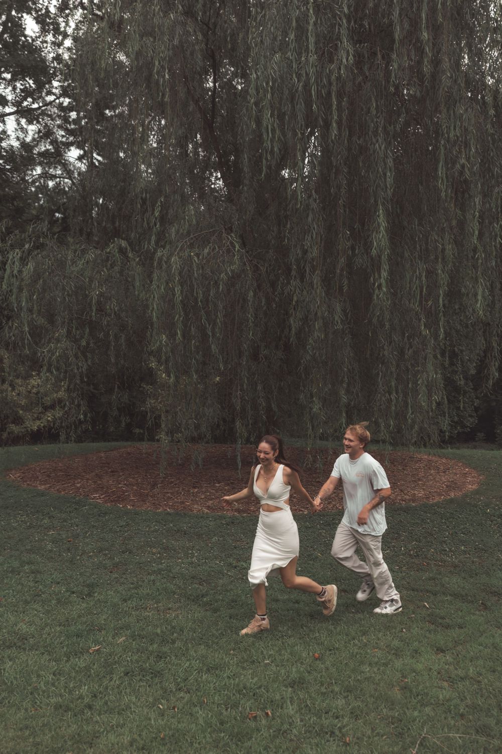A couple holding hands and running joyfully on green grass beneath a large weeping willow tree.