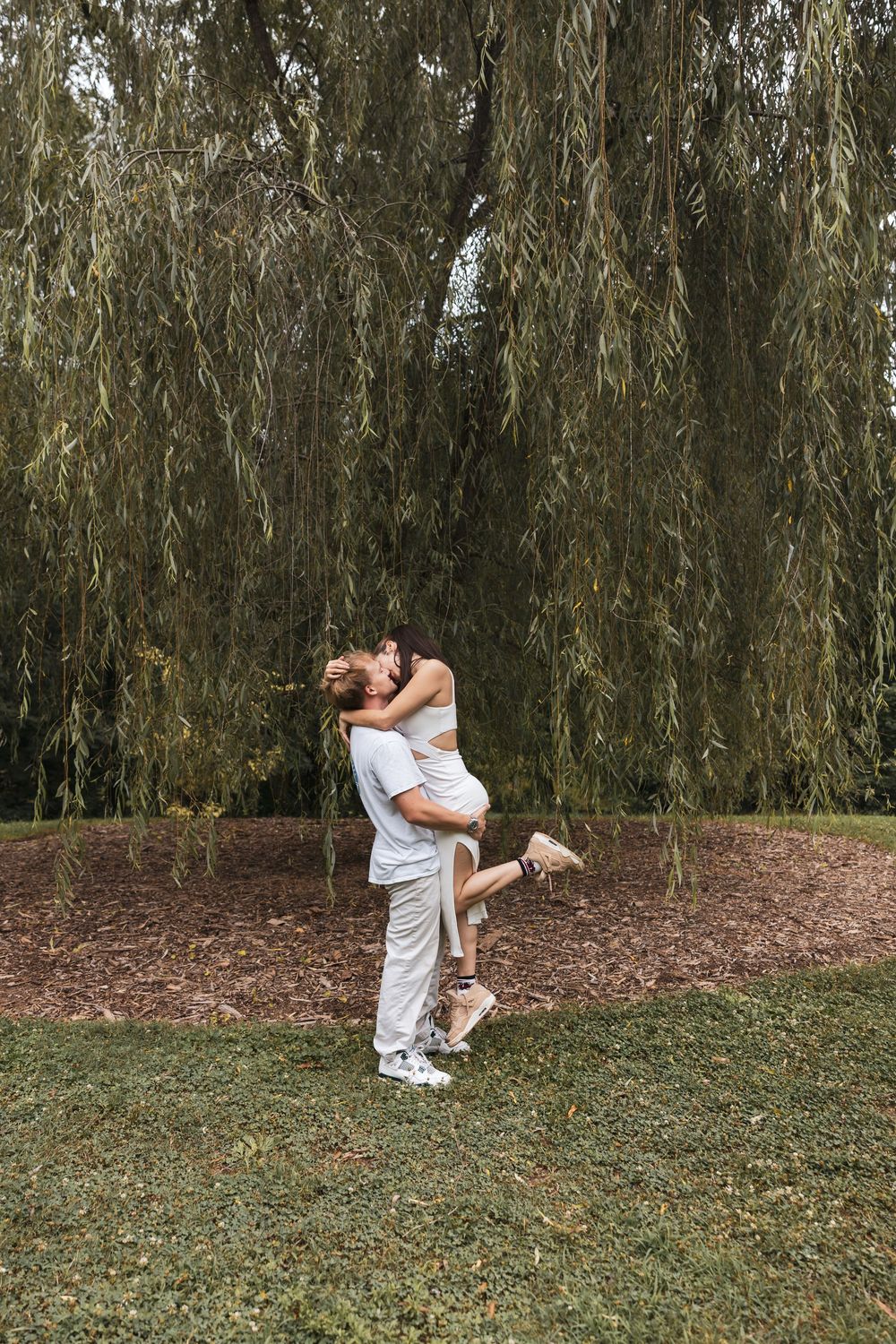 A couple shares a romantic kiss under a weeping willow tree, with the man lifting the woman in a lush green park.