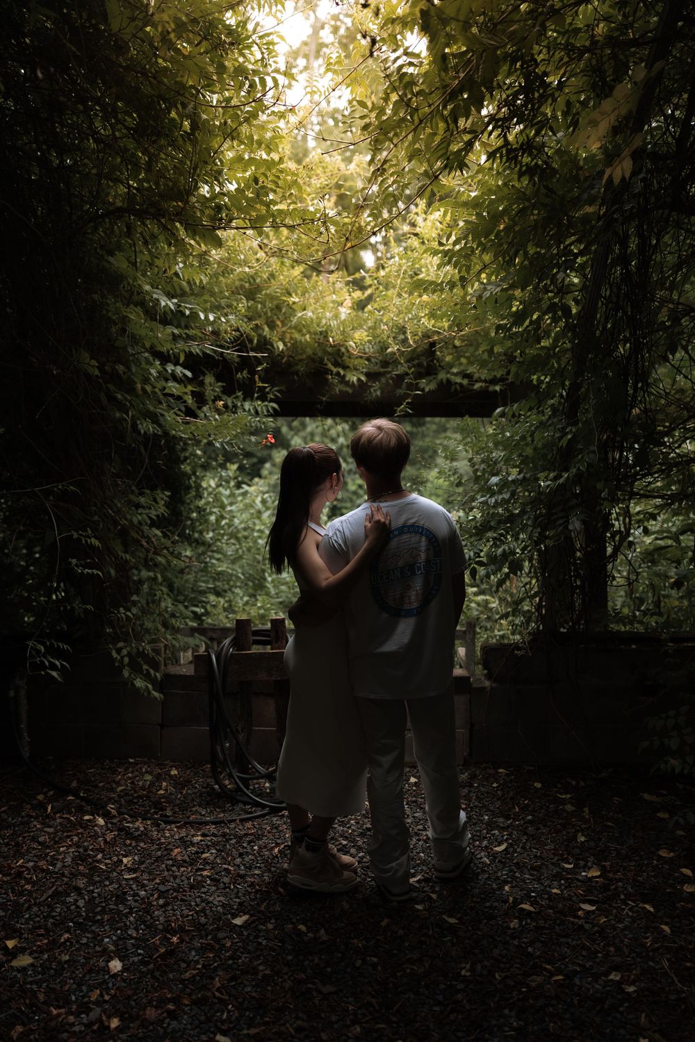 A couple embraces in a lush green forest tunnel, standing on a dark path surrounded by dense foliage and soft light.