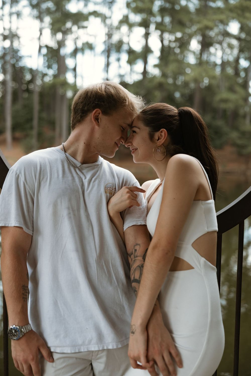 Romantic couple in white outfits sharing an intimate moment on a bridge surrounded by lush forest trees.