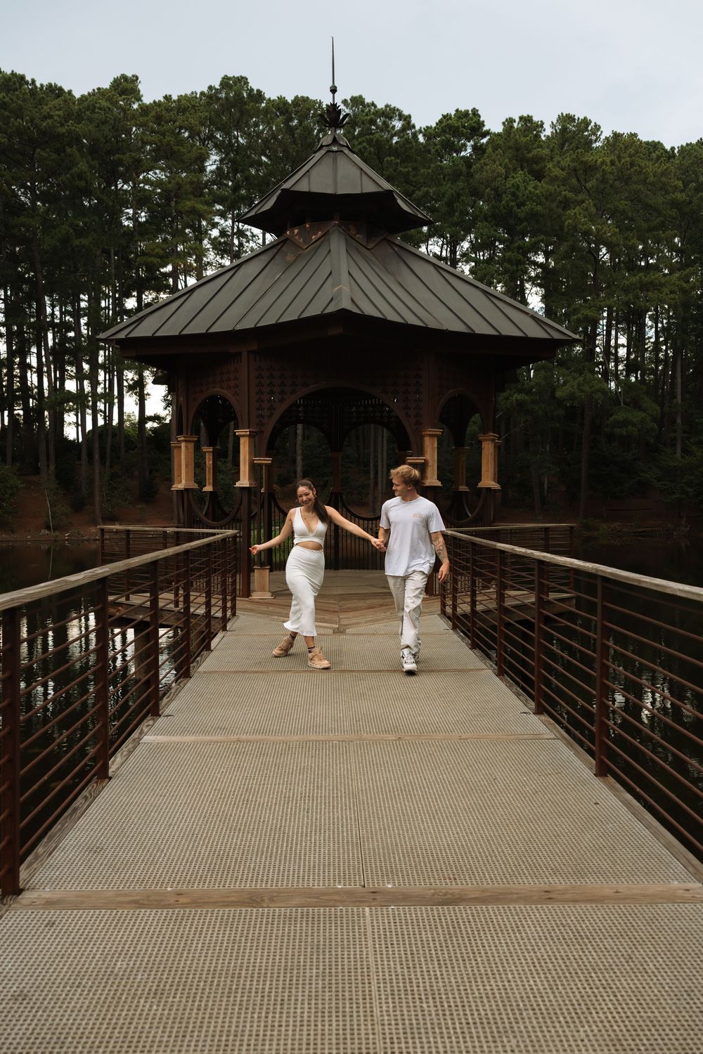 Couple holding hands walking on a bridge toward camera with an elegant gazebo and pine trees in the background.