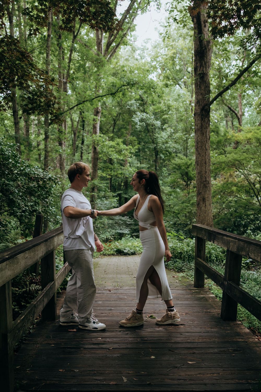 Couple holding hands on a wooden bridge surrounded by lush green forest trees in a romantic outdoor setting.