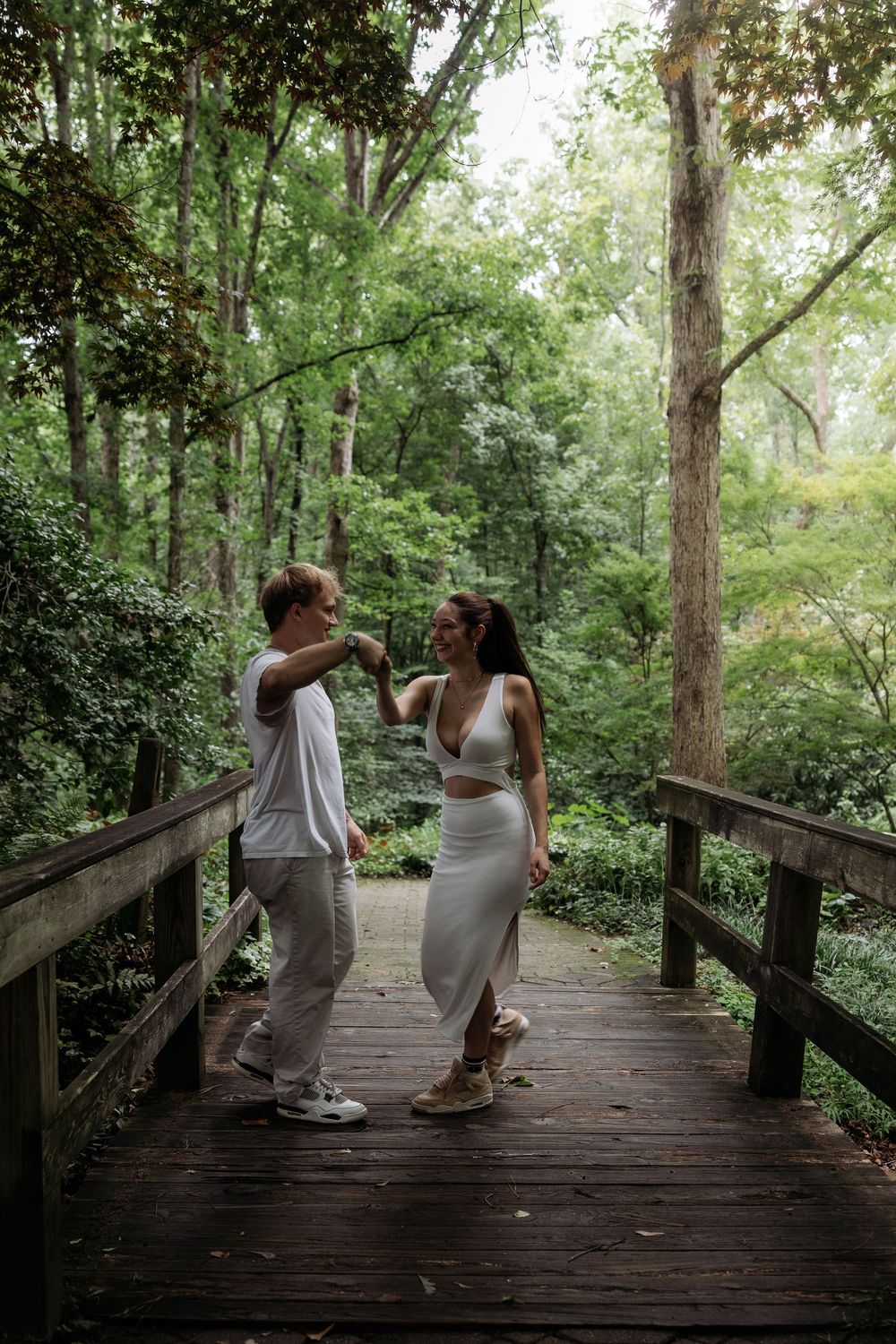 Couple dancing on a wooden bridge surrounded by lush green forest trees in a romantic outdoor setting.