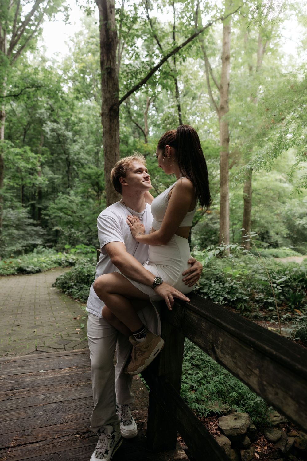 A happy couple shares an intimate moment on a wooden bridge surrounded by lush green forest trees.