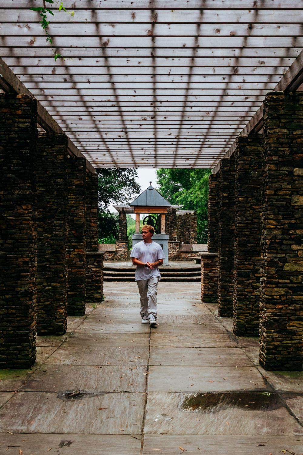Man walking through stone pergola pathway toward gazebo in lush garden park setting.