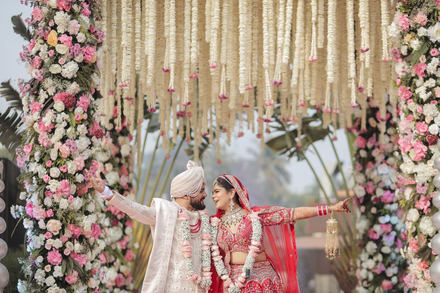Indian bride and groom in traditional wedding attire standing under floral mandap with pink and white flowers.
