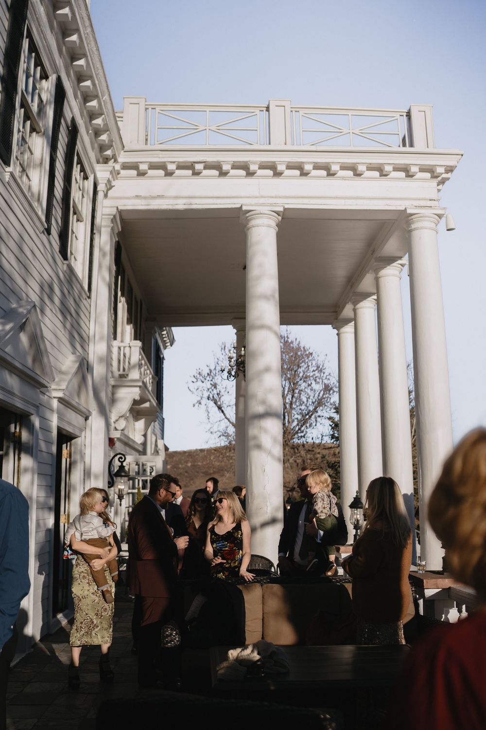 Guests mingle on the porch of a white colonial building with tall columns during an outdoor evening event.
