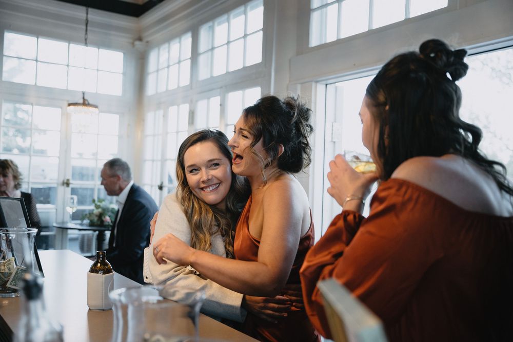 Three women laughing and embracing at a bright restaurant bar during a wedding reception celebration.