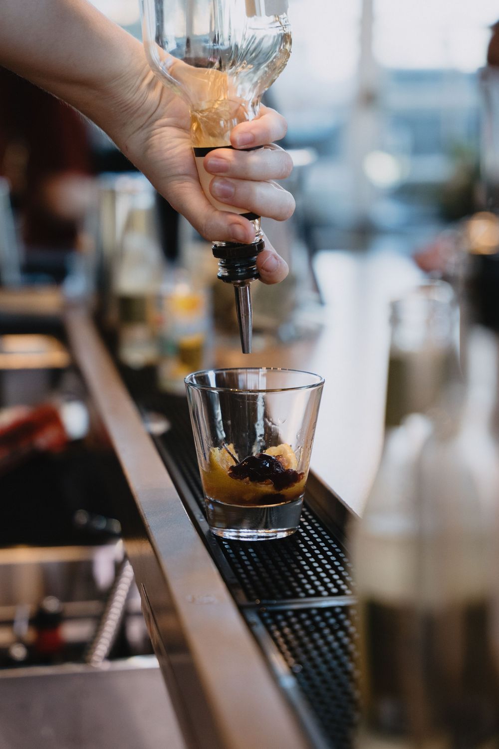 Bartender pouring dark liquor into a glass with ice at a bar counter using a bottle pourer.