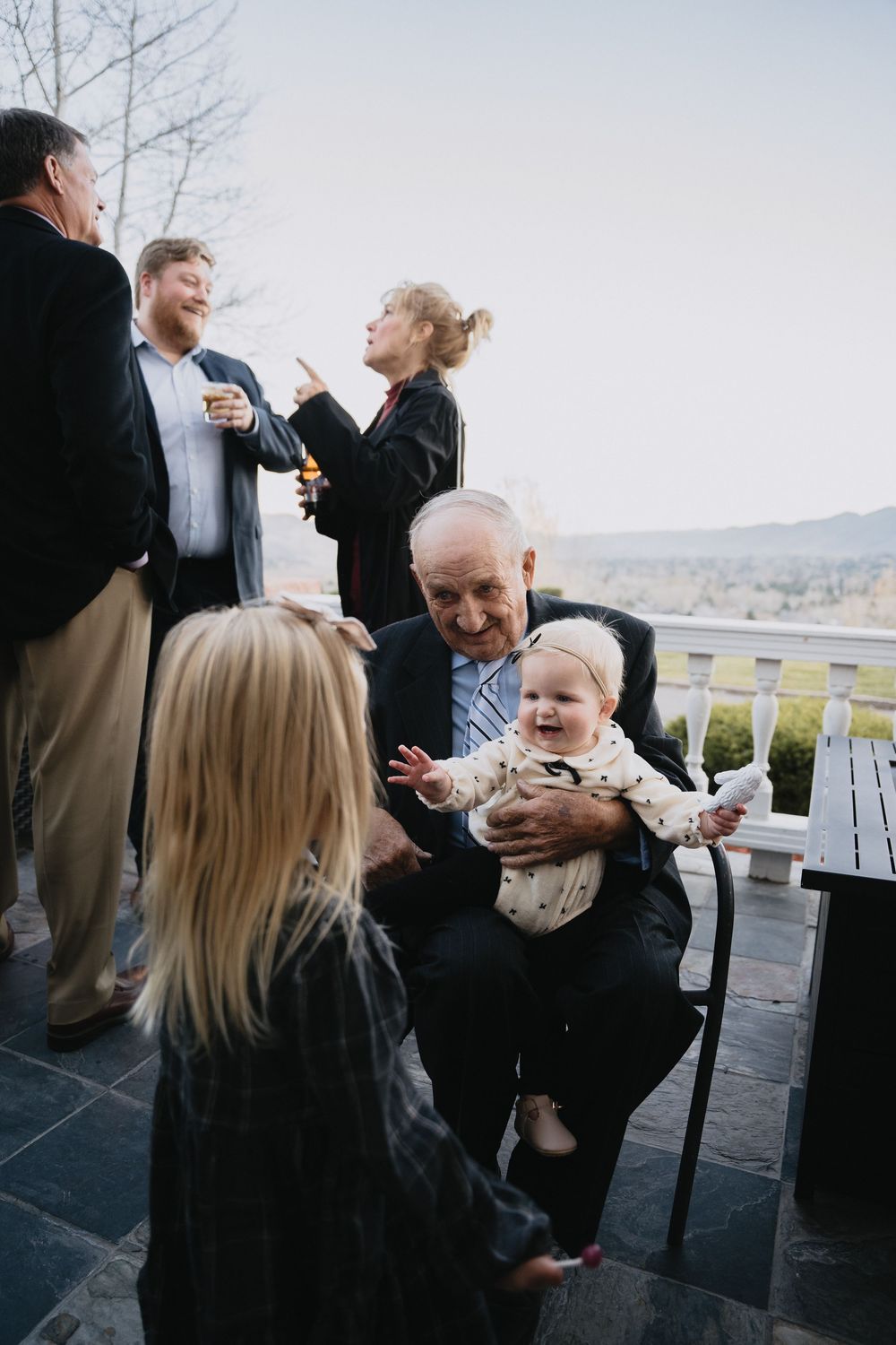 Elderly man holding a laughing baby on a porch, surrounded by family at an outdoor gathering.