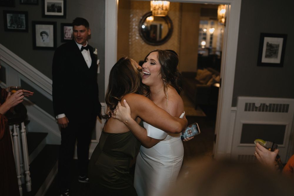 Bride in white gown hugging a guest in olive dress while groom stands near staircase at elegant wedding venue.