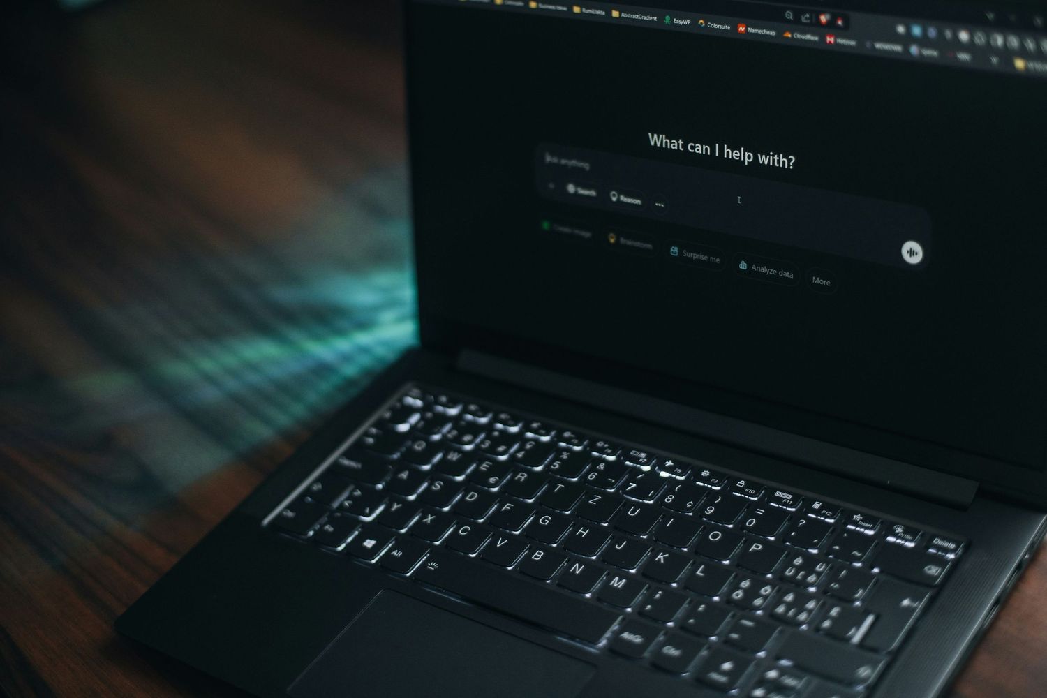 A dark laptop displaying an AI assistant interface with 'What can I help with?' on screen, placed on a wooden desk.