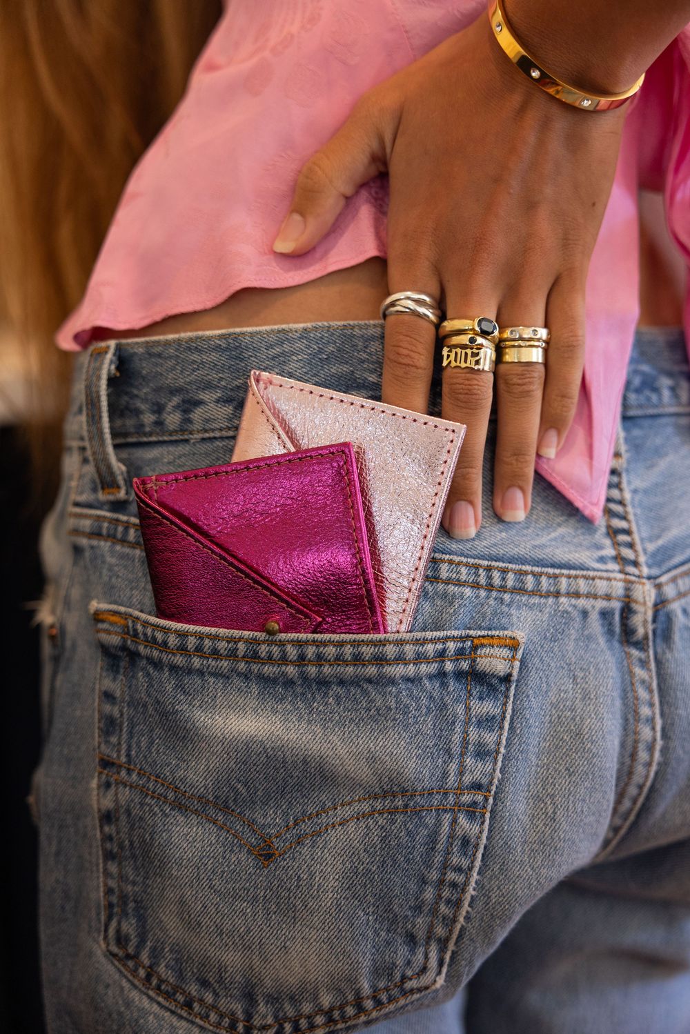 Woman in pink top and jeans with gold rings holding pink metallic envelope card wallets in back pocket.